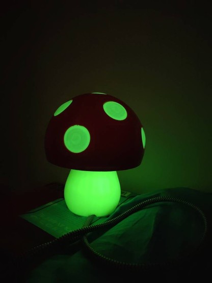 Eine Lampe in rot gehalten mit grünlich leuchtendem Lampenfuß und leuchtenden Flecken im Lampenschirm. Die Lampe sieht aus wie ein grün leuchtender Fliegenpilz.